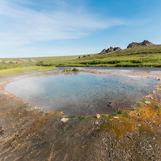 A shallow hot spring in a remote landscape is surrounded by rolling hills. 