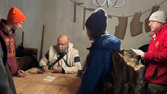 Young men play a card game with an older man dressed in historic costume. 