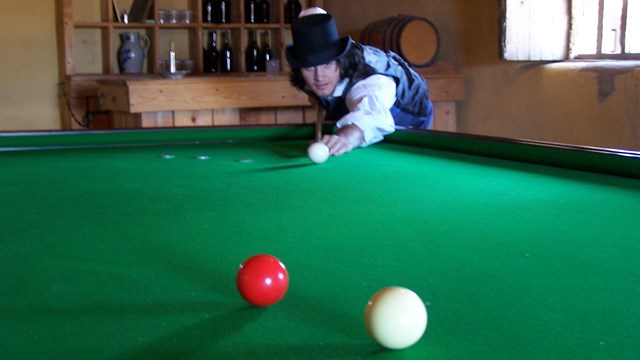 A man in historic costume leans over a pool table with a cue in hand. 