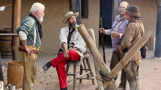 Four men dressed in 19th century historic costume sit and stand near a smoldering fire. 