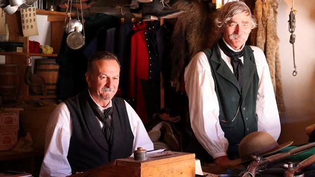 Men in historic costume stand behind a wooden sales counter. 