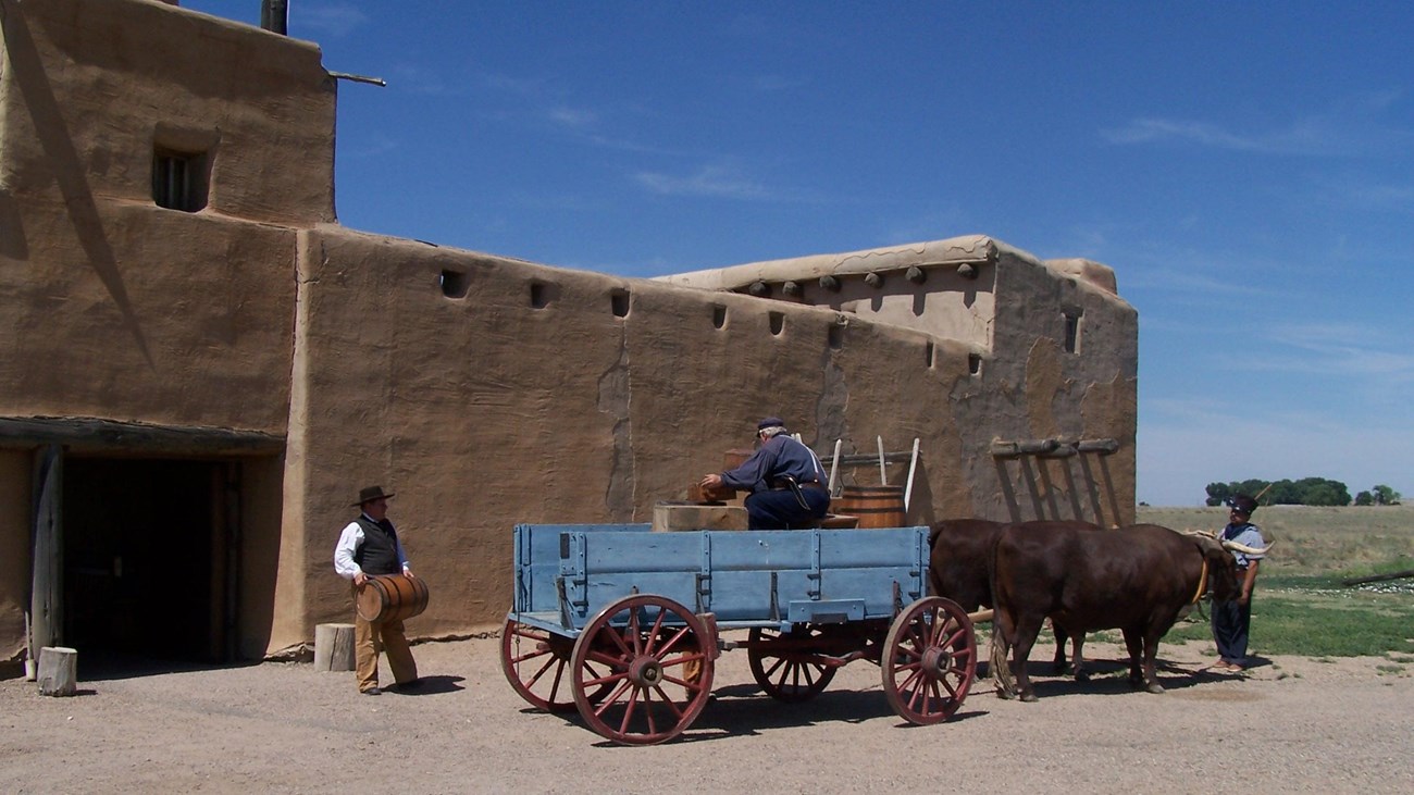 Men unload a wagon pulled by oxen in front of an adobe fort
