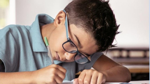 boy with glasses and short-sleeve shirt sitting at desk with pencils writing on a page 