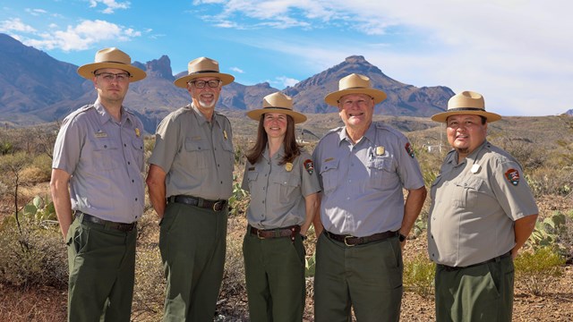 Five people wearing park service uniforms stand in a line in front of the Chisos Mountains.