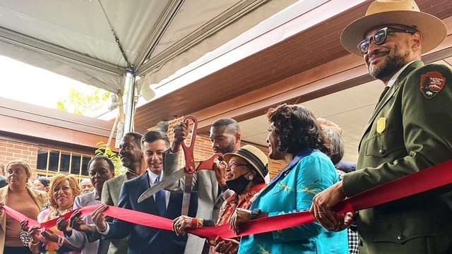 A line of people stands behind a red ribbon. A man in a grey suit holds a pair of scissors. 