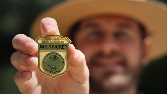 A park ranger holding a gold plastic badge that reads "junior ranger; big thicket national preserve"