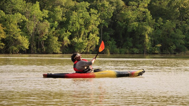 a person in a black-red-yellow kayak paddling along a wide waterway and smiling at the camera.