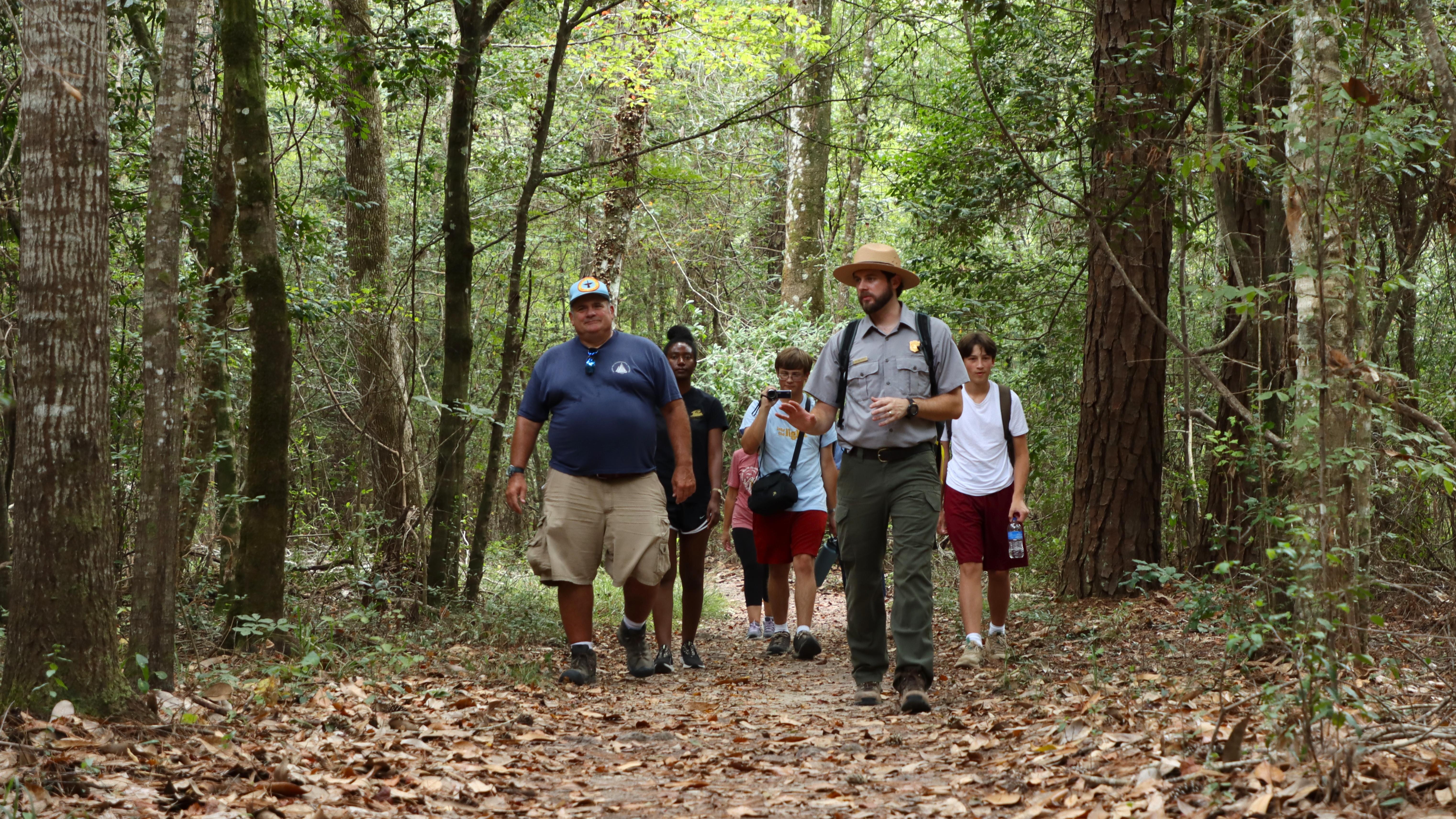 A park ranger leading a group of hikers along a leaf-covered trail through thick green forest.