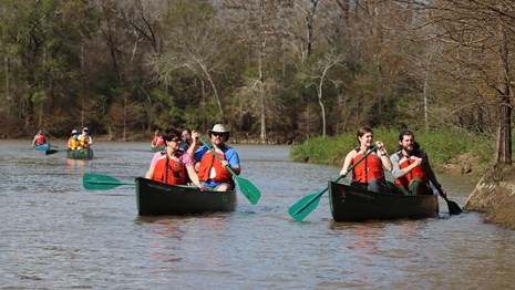 A group of canoeists paddling in canoes on a wide waterway, while wearing orange life jackets.