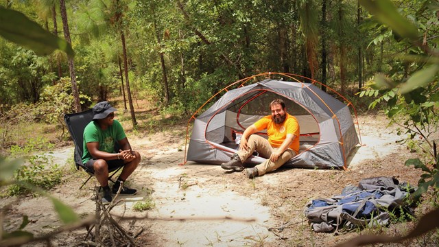 2 people at a campsite. 1 person sits in a folding chair while another is sitting in a small tent.