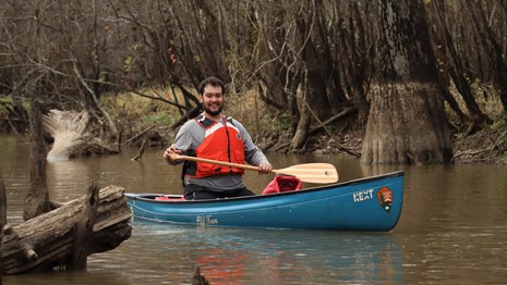 A paddler in orange life vest with wooden paddle and blue canoe on the water.