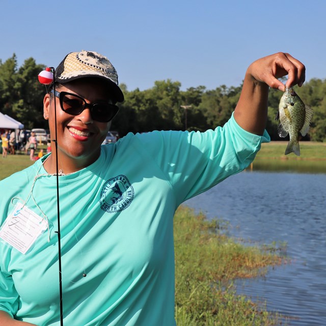 smiling woman holding a small green-gray fish that she caught from a nearby pond.