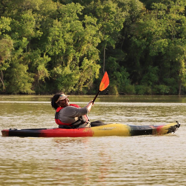 a person in a black-red-yellow kayak paddling along a wide waterway and smiling at the camera.