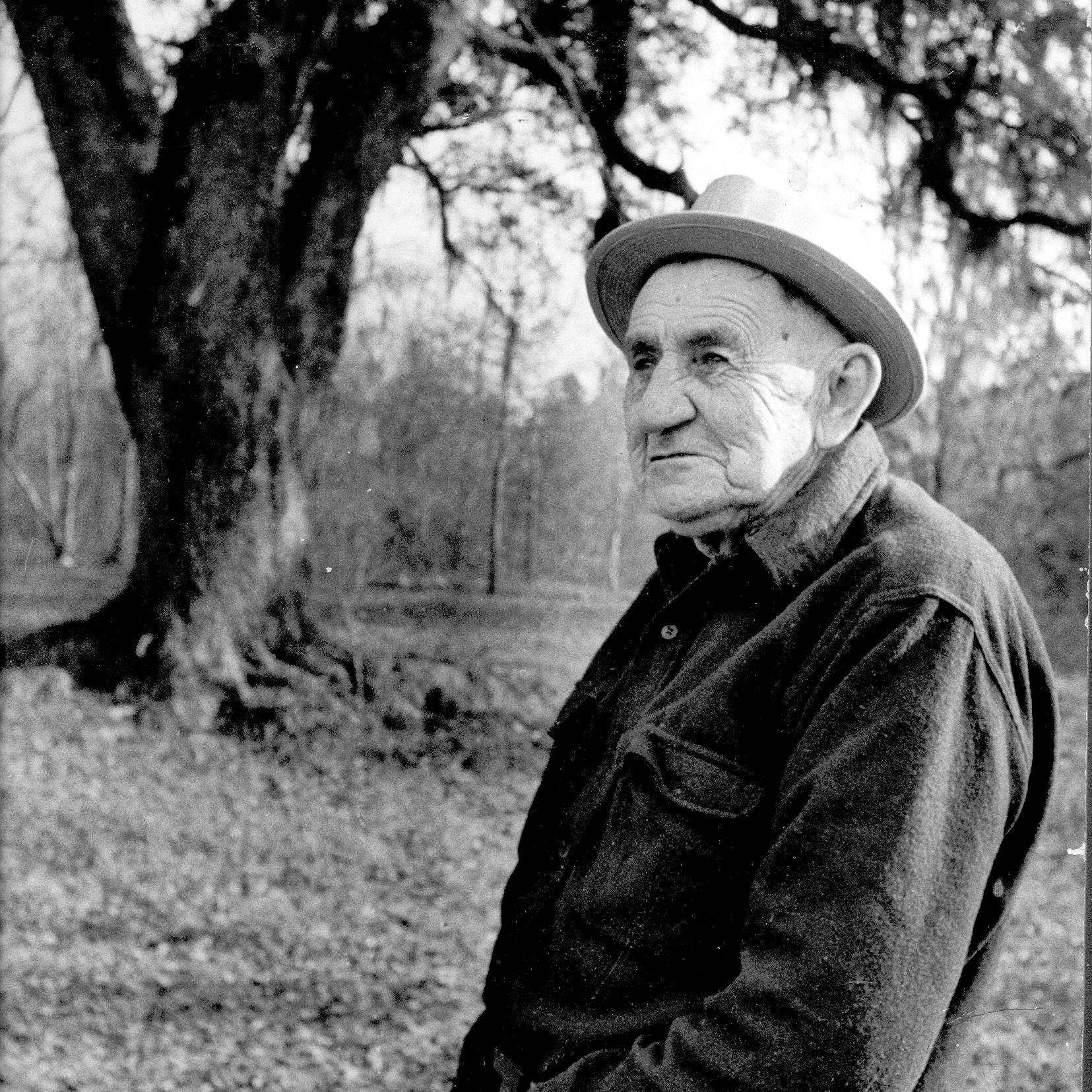 Black and white photo of an old man standing near a large oak tree.