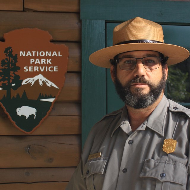 ranger standing in front wooden building and NPS arrowhead logo