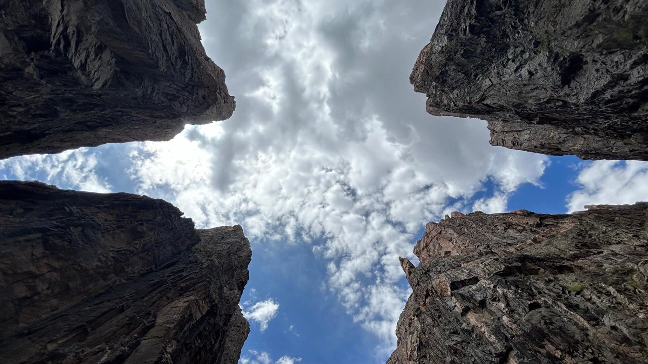 Four dark canyon walls with a blue and cloudy sky. The perspective is looking up at the walls.