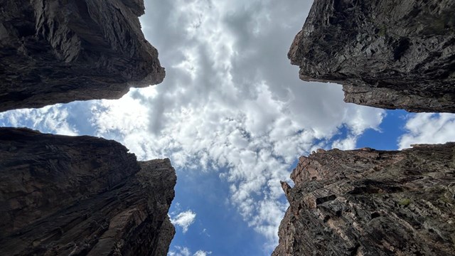 Four dark canyon walls with a blue and cloudy sky. The perspective is looking up at the walls.