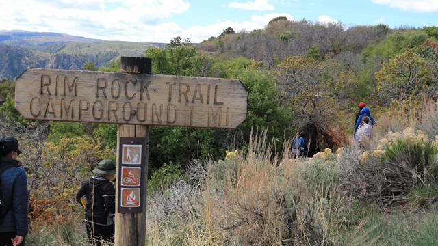 Adults and children with jackets and backpacks hike along a trail. Wooden trail sign in foreground.