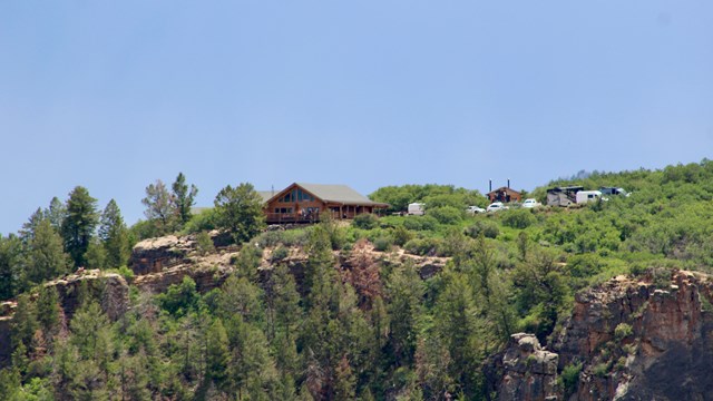 A log cabin style building with a parking lot sits near a forested canyon rim. Blue sky above it.