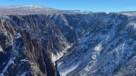 Snow covered steep canyon walls with dark river flowing at the bottom.