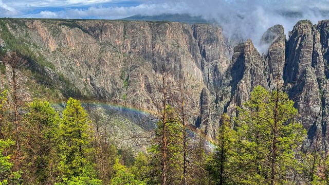 Steep grey-black canyon walls with coniferous trees in the foreground. A rainbow stretches across