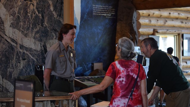 A person in a park service uniform stands behind a desk. Two visitors are asking them questions