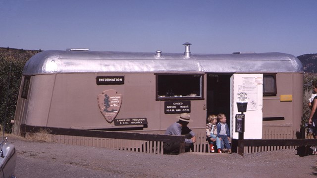Park ranger squats down beside two children in front of a beige and metal information trailer.