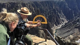 Ranger holding a protractor standing in front of the black canyon being filmed by another ranger.