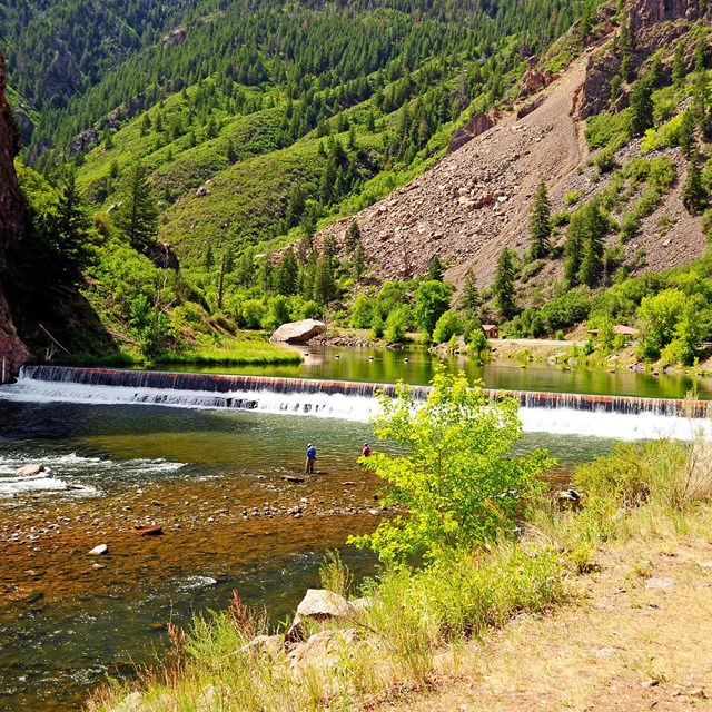 fisherman wading in the river in front of a short waterfall in the canyon