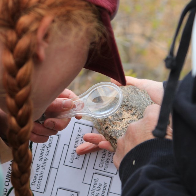 A child holds a magnifying glass up to a rock in front of a clipboard