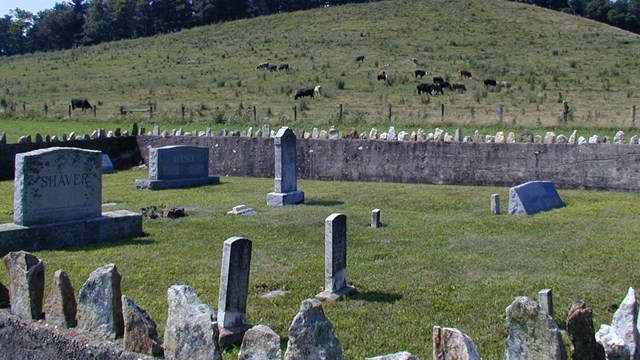 stone headstones scattered throughout a plot of grass surrounded by a hand laid stone fence
