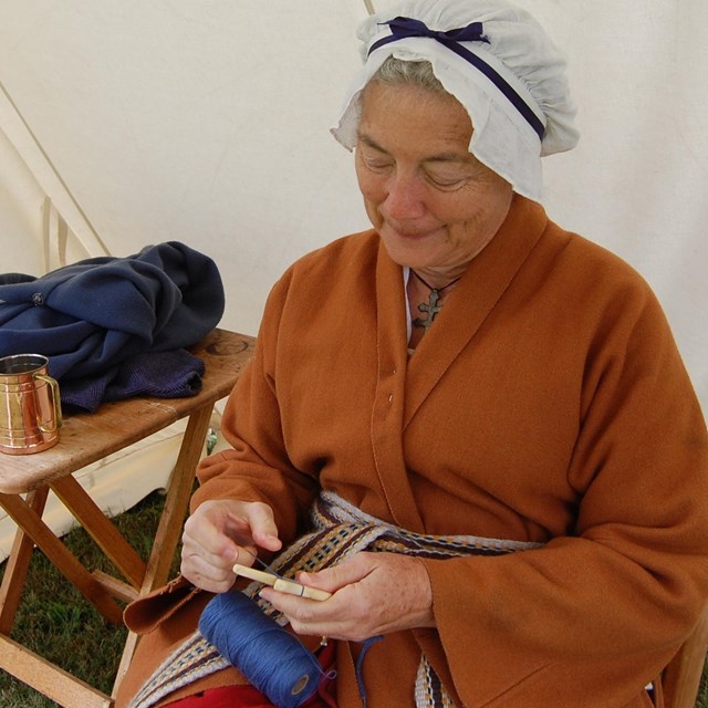 A woman in Revolutionary War Era clothing making a cord with a lucet tool.