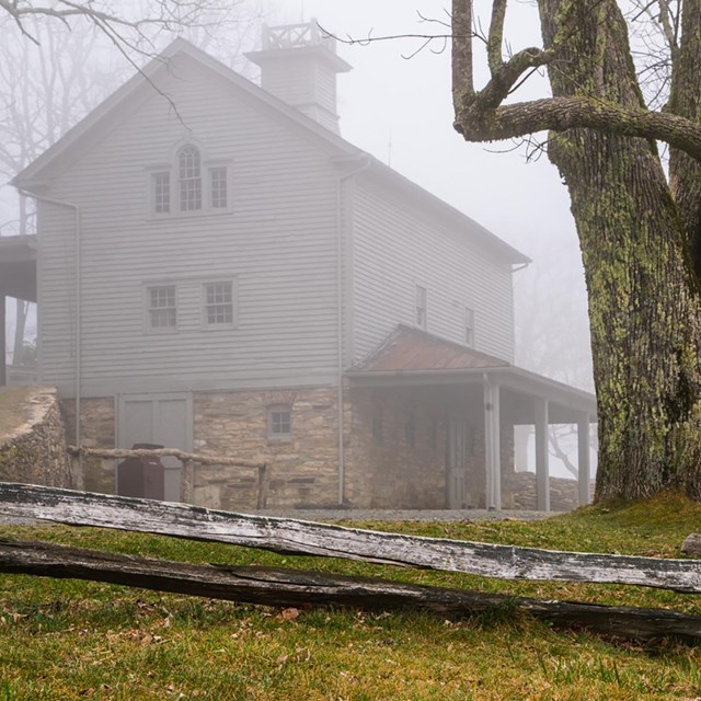 An old, wooden building surrounded by large trees and a split-rail fence