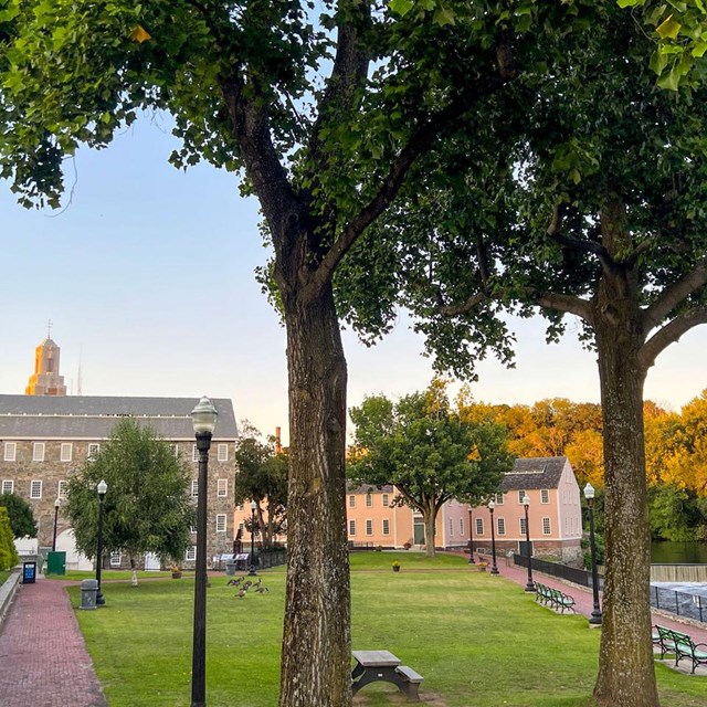 A tree-lined park with brick pathways, benches, and a river, with historic buildings in the backgrou