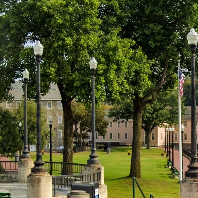 A riverside park with a monument, lampposts, and historic mill buildings in the background.
