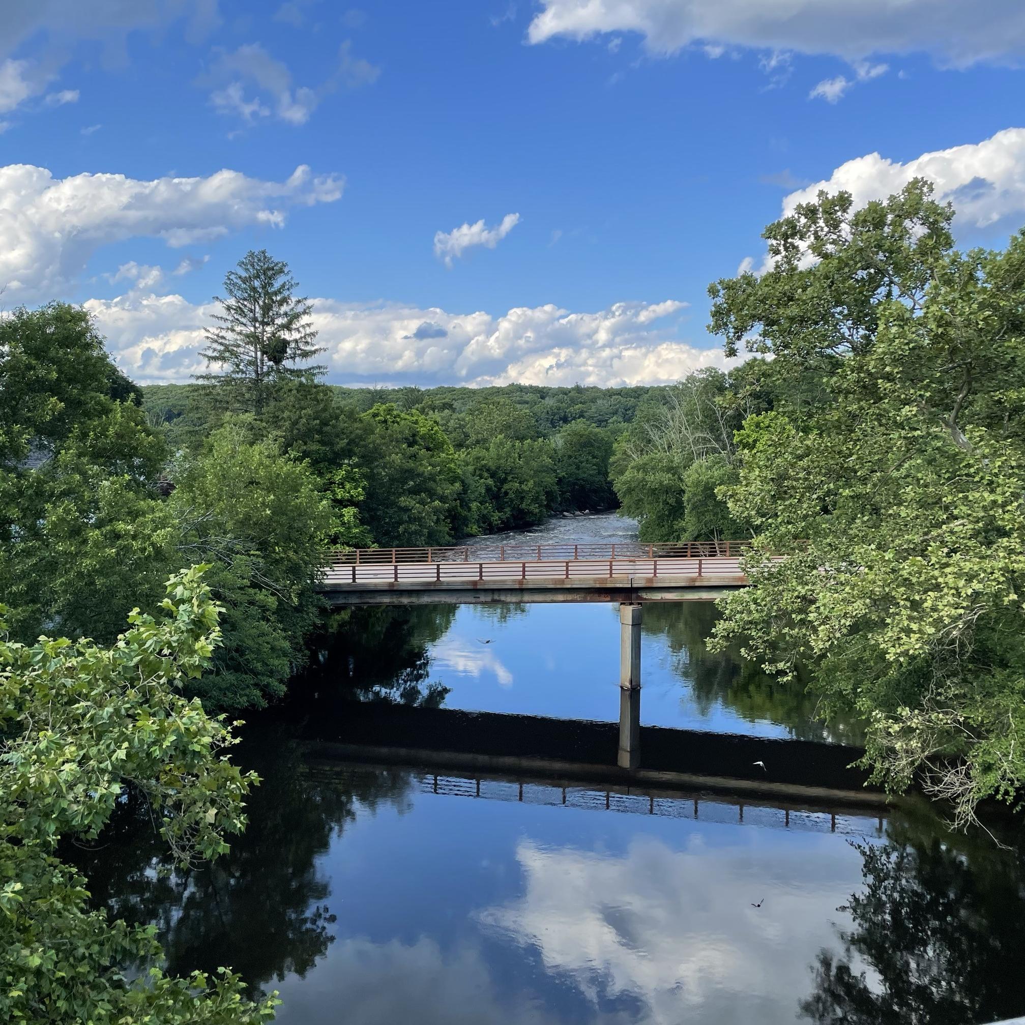 River with bridge crossing it with trees on either side and reflection of clouds in water