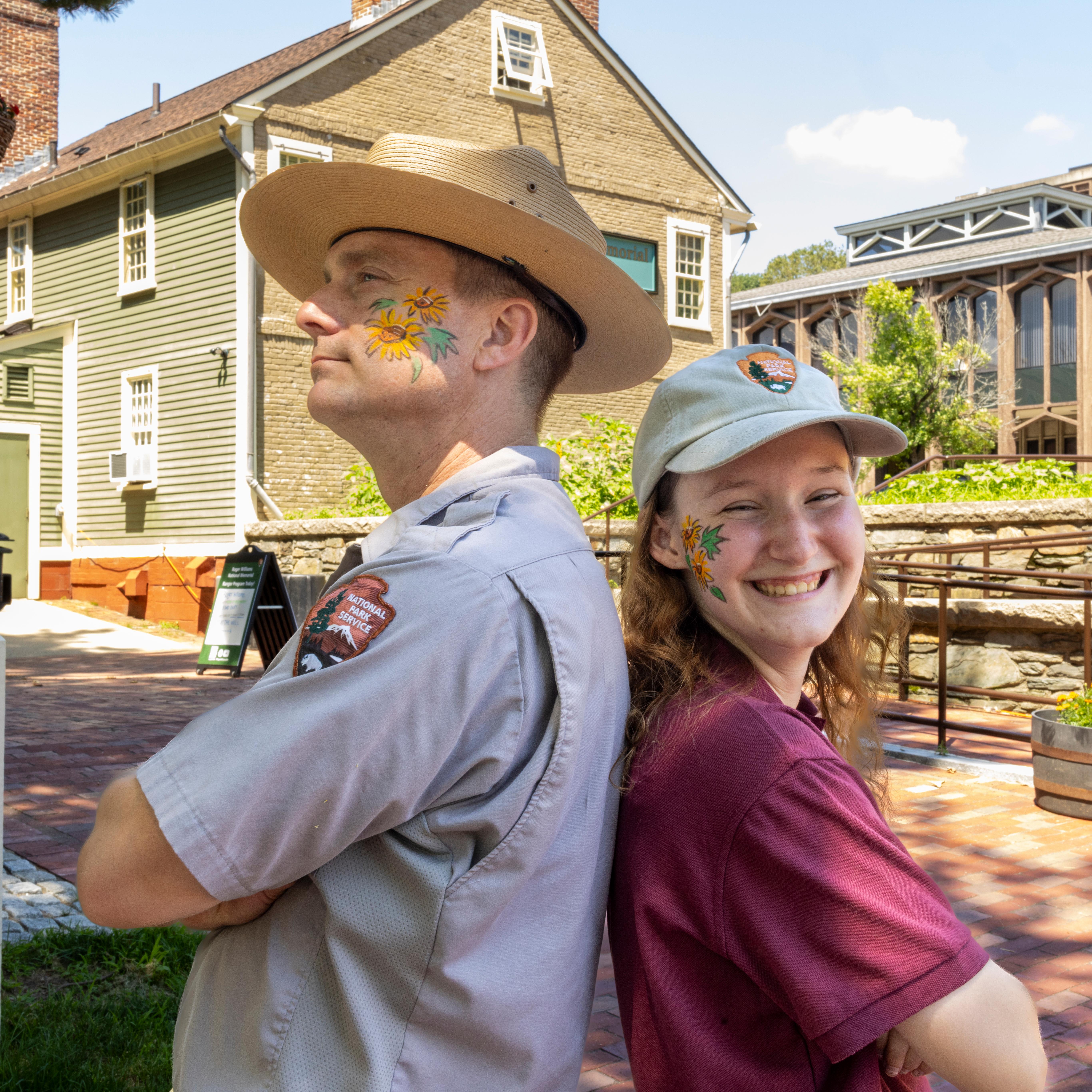 Park Ranger and Intern stand back to back in front of Visitor Center with sunflowers painted on face