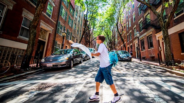A boy carrying a white flag crosses a tree-lined street.