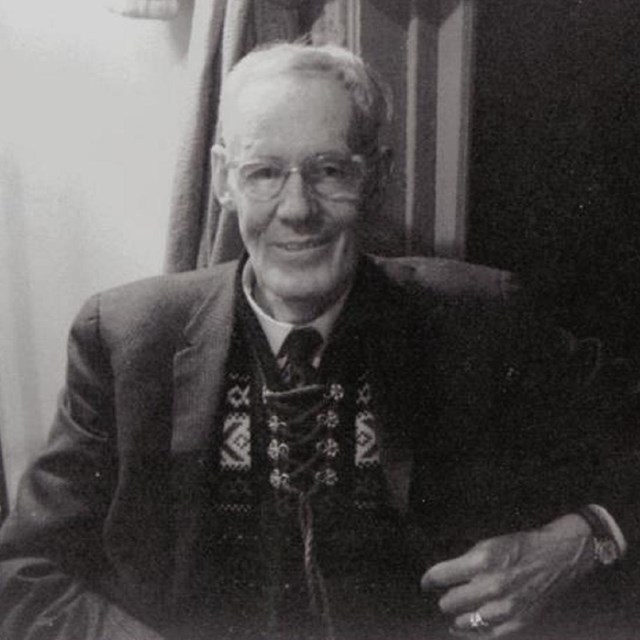 older white man sitting in a chair. He wears a short white beard and round glasses and wears a suit.