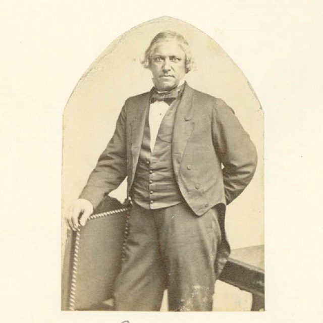 Historical photograph of a man standing next to a chair with his right hand resting on chair back.