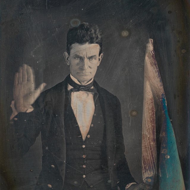 A middle-aged man (John Brown) raising one hand and holding a standing flag in the other. 