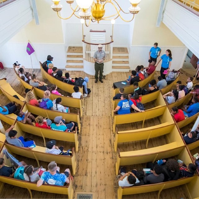 2nd floor view in African Meeting House looking onto students seated in pews with ranger speaking