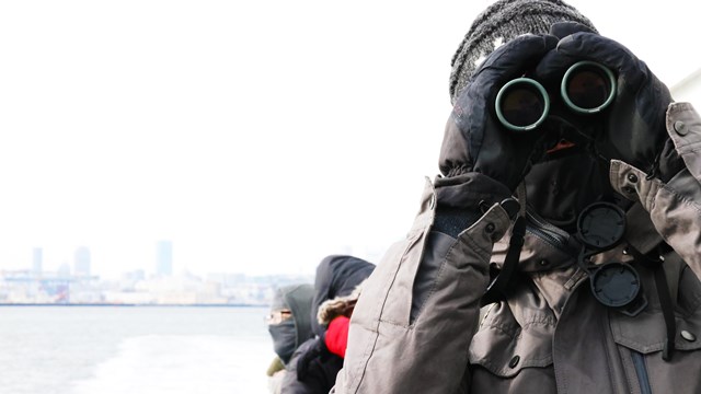person in winter gear on a boat looking at the camera via binoculars 