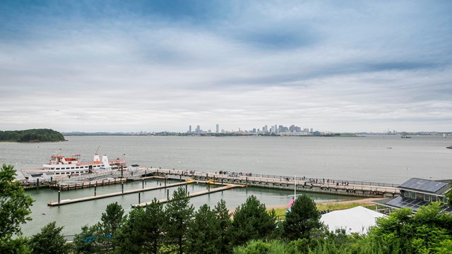 view from island drumlin looking down on dock with a ferry. Background is Boston Harbor & skyline. 
