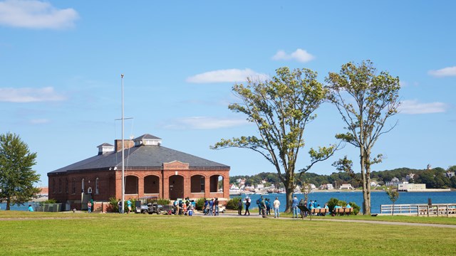 green lawn with a couple tall trees and a brick one story building with water in background.