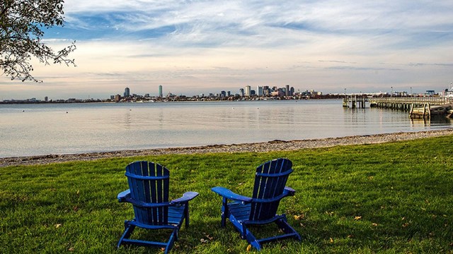 two blue Adirondack chairs on grassy knoll leading to beach. Dock to the right & city in distance. 