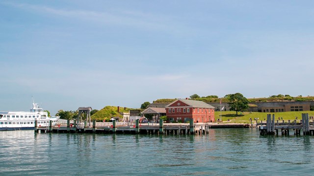 View of Georges Island from the water. See docks, a ferry, and visitor center. Fort in background.