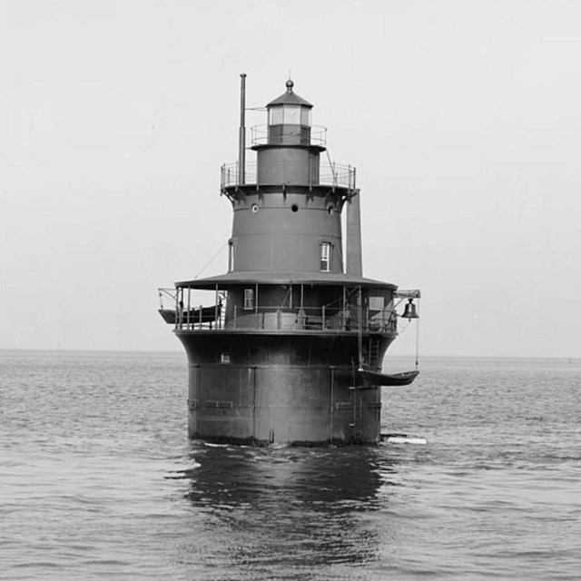 Round, short, metal buoy lighthouse next to a small cropping of rocks in water. 