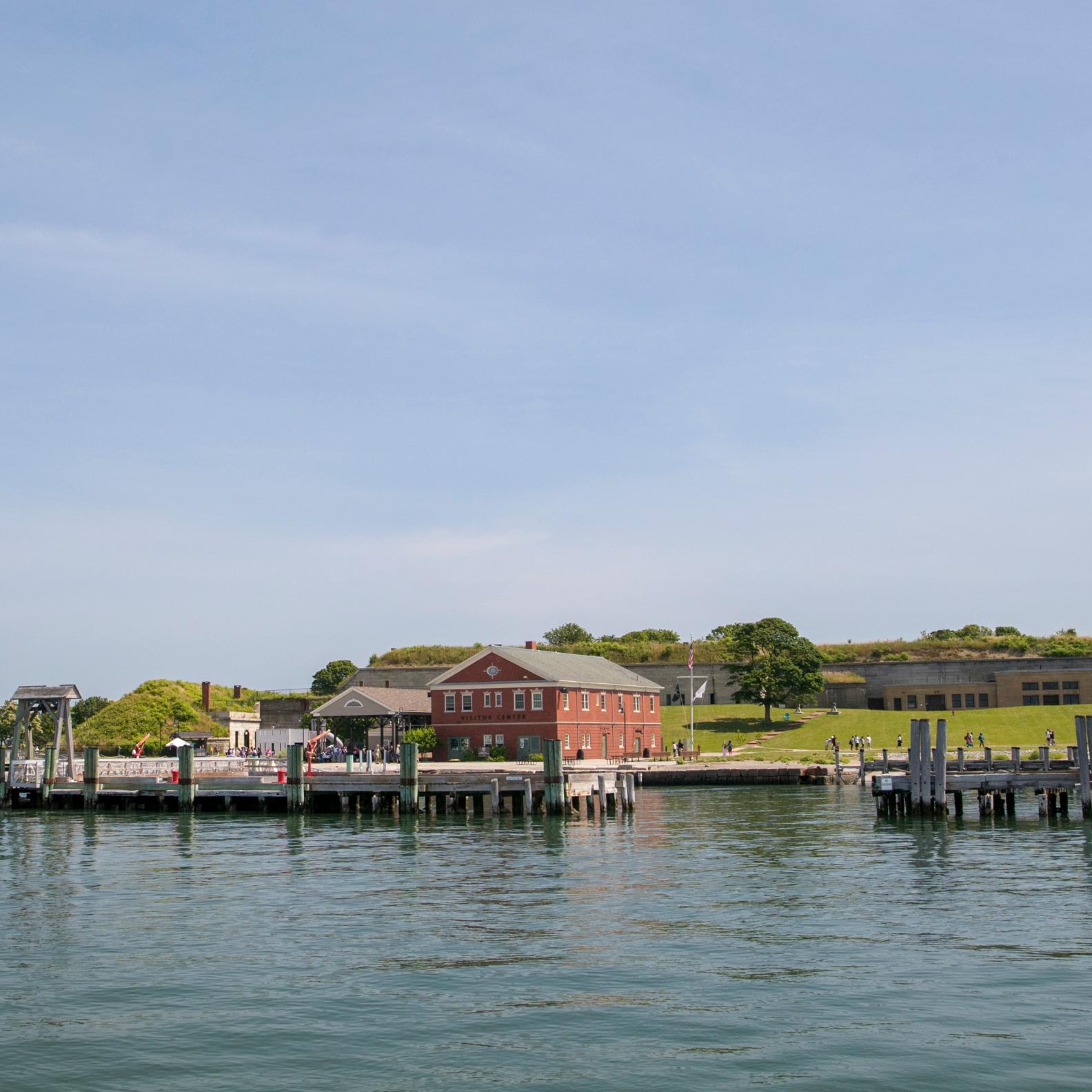 View of Georges Island from the water. See docks, a ferry, and visitor center. Fort in background.