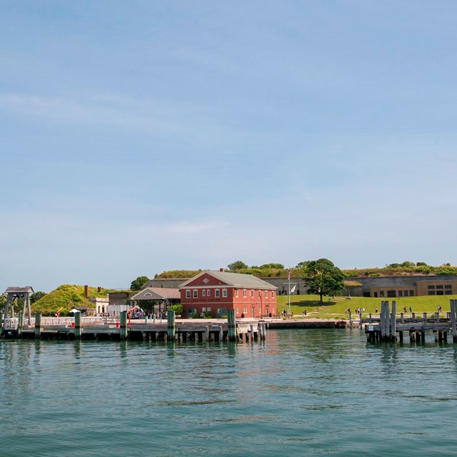 View of Georges Island from the water. See docks, a ferry, and visitor center. Fort in background.